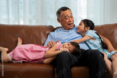 Happy Asian senior grandfather sits on a couch with his granddaughter and plays with his granddaughter in the living room at home, The Concept of family having fun in their house