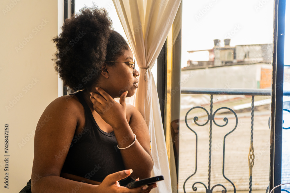 joven mujer afroamericana sentada al lado de la ventana de la casa mirando distraída - obrazy, fototapety, plakaty 