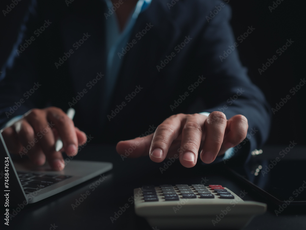 Businessman in black suit working on laptop computer and tablet and calculator, Hand Hands touch on calculator at office with dark background, Online working, Close up, Copy space. Businessman in black suit working on laptop computer and tablet and calculator, Hand Hands touch on calculator at office with dark background, Online working, Close up, Copy space.