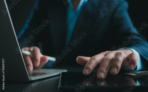 Businessman in black suit working on laptop computer and tablet, Hand Hands touch on tablet at office with dark background, Online working, Close up, Copy space.