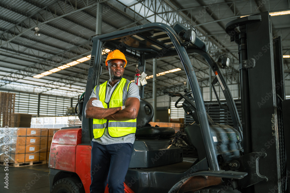 African forklift drivers focused on carefully transporting stock from shelves of a large warehouse wearing a helmet and vest looking toward goods