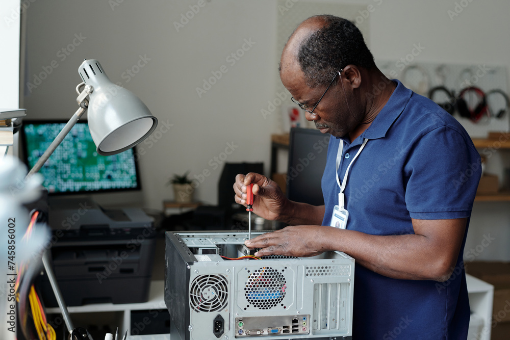 Side view of mature professional technician with screwdriver in hand fixing details of computer hardware while standing by workplace