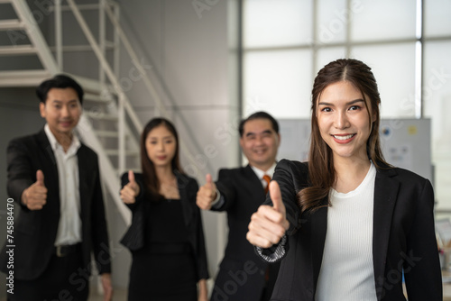 Beautiful businesswoman gives a thumbs up to the camera with an attractive smile. She stands with her team at the office. Businessman concept.	