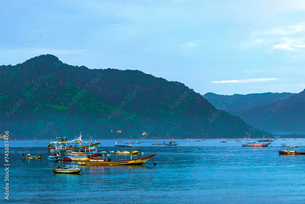 A photo of colorful boats anchored near a beach at sunrise in Trenggalek, Indonesia, with mountains in the distance.