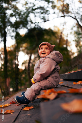 A small beautiful baby in a warm overalls sits on a wooden ladder. A walk on the street among the yellow leaves.