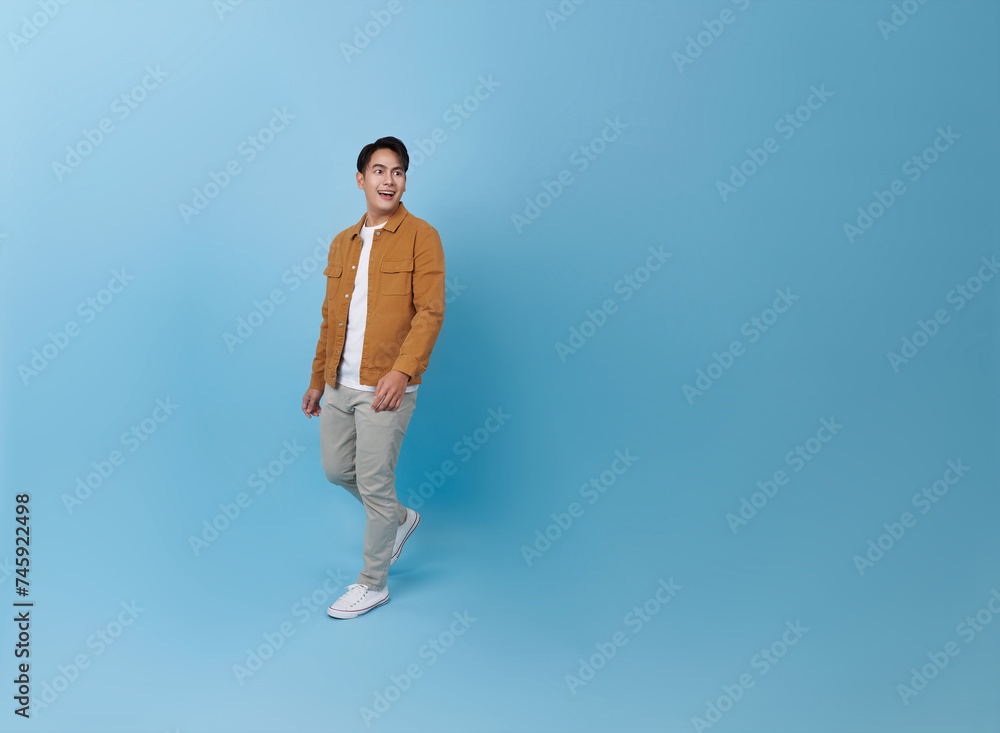 Full body smiling happy young Asian teen man walking going strolling and looking sideways isolated on blue background studio portrait.