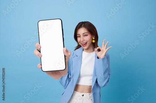 Beautiful Asian teen woman holding smartphone mockup of blank screen and shows ok sign isolated on blue background.