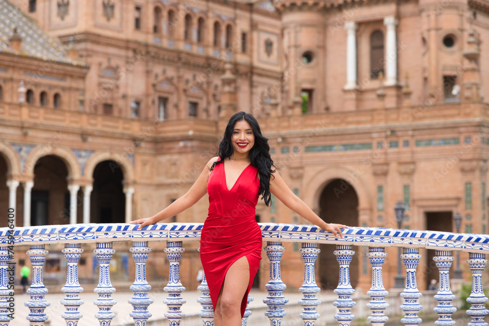 Young and beautiful brunette and latin woman, dressed in short red dress and red shoes visits the famous square in seville, spain. Woman leaning on the railing of a tiled bridge and is happy. Travel.