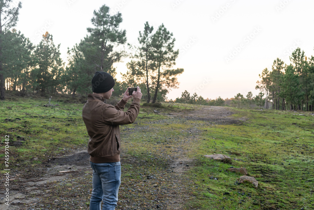 Man standing with a fur jacket and wool hat in the forest taking a photo with his mobile phone Man standing with a fur jacket and wool hat in the forest taking a photo with his mobile phone