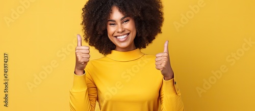 A cheerful African American woman with a yellow shirt is laughing as she gives a thumbs up gesture. She is isolated on a yellow background, expressing positivity and approval.