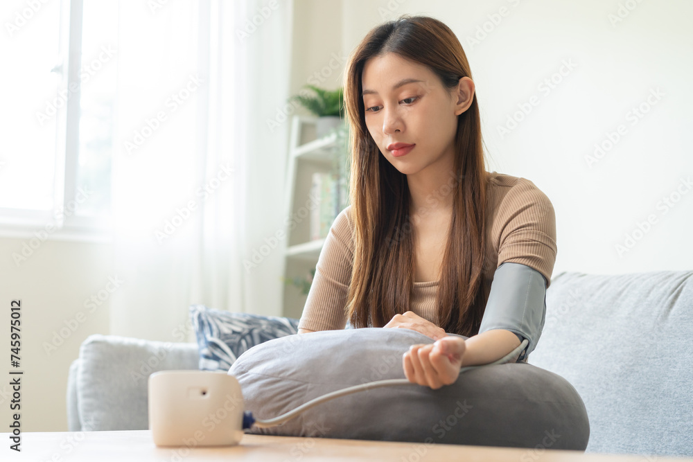 Hypotension, asian young woman sitting on couch checking blood high pressure and heart rate with digital pressure monitor machine making self check up on her arm at home. Healthcare and medical.
