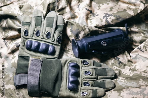 Tactical military gloves and Thermal imaging army sight on the khaki camouflage uniform. Black Thermographic camera, close up. Military industry, war, global arms trade.