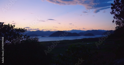 A picturesque evening view framed by silhouetted foliage, overlooking a lake with mountain ranges under a twilight sunset sky