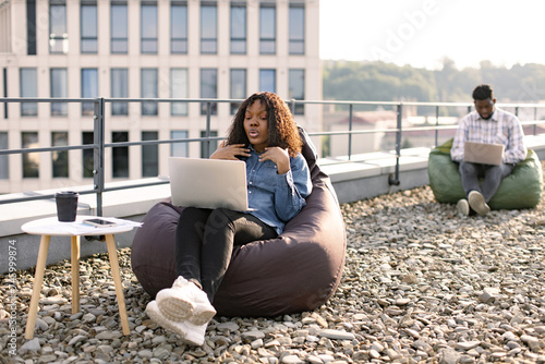 Happy African American female person in casual clothing gesture at laptop camera on flat roof of building, while her male colleague working in blurred background. © sofiko14