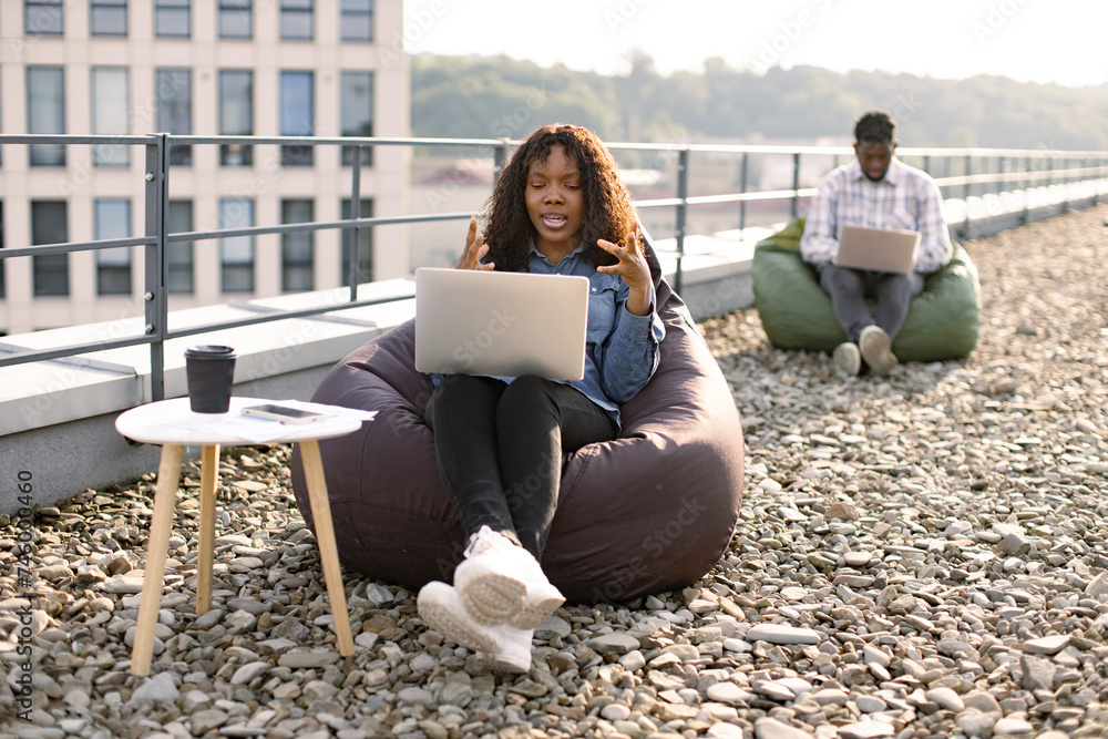 Efficient African American female freelancer beginning video conference via internet connection outdoors, while her male colleague working in blurred background.
