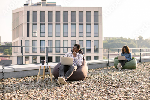 Smiling male using laptop outdoors sitting in bag chair, female colleague works in background. Modern lifestyle, connection, business, communication, web chat, social media, video conference concept.