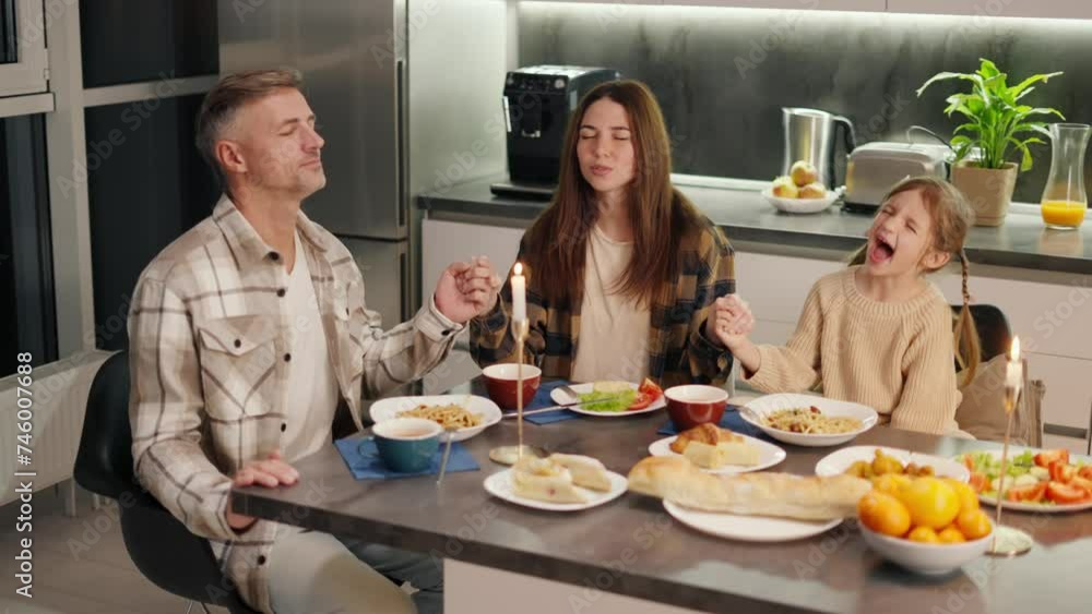 Happy middle aged gray haired man with his brunette wife and little girl praying and giving thanks for food before starting dinner at the dining table in the kitchen Happy middle aged gray haired man with his brunette wife and little girl praying and giving thanks for food before starting dinner at the dining table in the kitchen