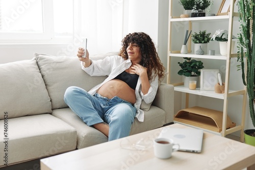 Pregnant woman blogger sits on the couch at home and takes pictures of herself on the phone, selfie and video call, consultation with the doctor online, pregnancy management