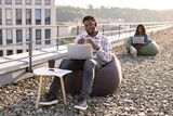Focused African male wearing denim shirt and headset holding online conference over portable computer on rooftop, female colleague working on blurred background.