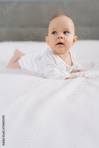 Vertical shot of pretty infant baby in white bodysuit lying on stomach and happily waves arms and legs with smile. Portrait of nice newborn child learning to hold head on comfortable blanket at home.