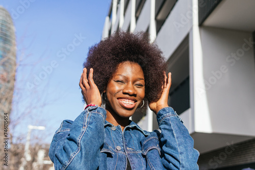 An exuberant African American woman touches her hair and smiles widely, basking in the sunlight against a modern city backdrop photo