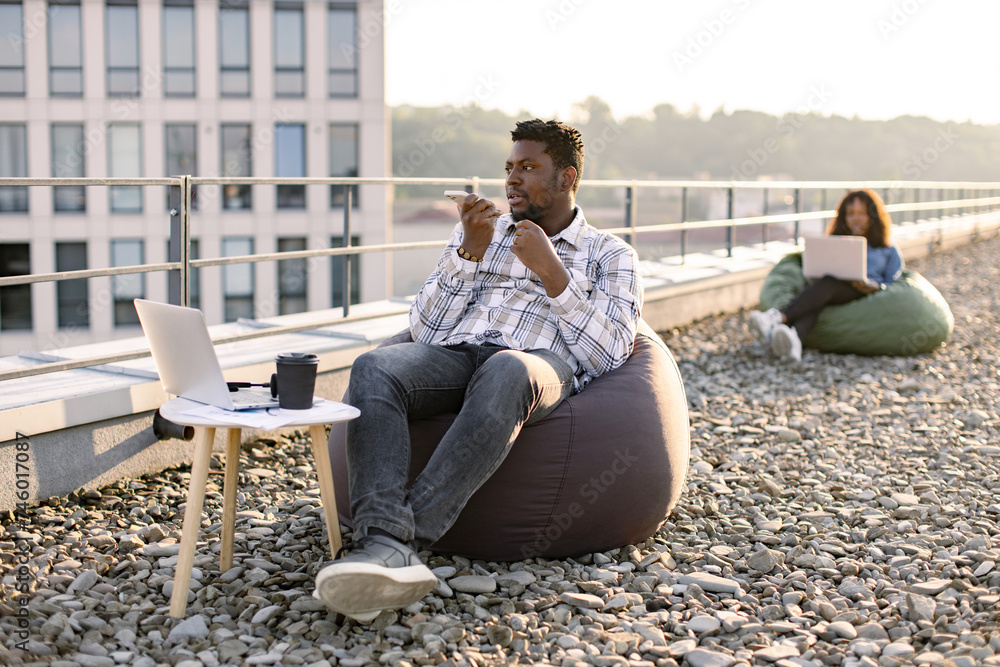 Young businessman wearing checkered shirt while holding smartphone and recording audio message, sitting in pouf chair outdoors while female colleague working on blurred background. Young businessman wearing checkered shirt while holding smartphone and recording audio message, sitting in pouf chair outdoors while female colleague working on blurred background.