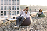 Carefree African American man in casual wear making phone call while relaxing in bean bag on flat roof while female colleague working on blurred background.
