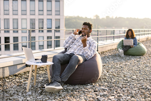 Carefree African American man in casual wear making phone call while relaxing in bean bag on flat roof while female colleague working on blurred background. © sofiko14