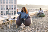 Joyful African woman in casual denim shirt using phone while recording video for her vlog on panoramic terrace, while male colleague working on blurred background.