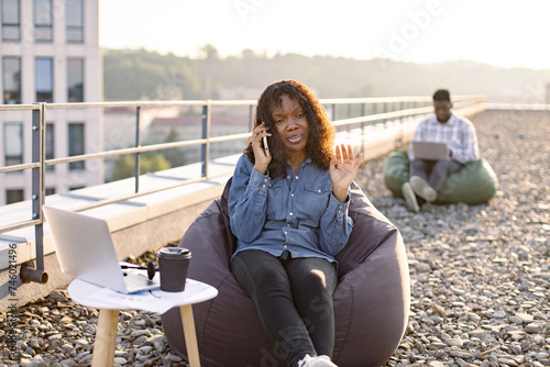 Beautiful adult African American female discussing agreement using smartphone while doing outdoor office work, businessman colleague working on blurred background.