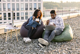 African man and woman working on laptop while sitting at rooftop workplace of office during break. Young coworkers eating healthy salad at lunch, sitting at cozy bean bag outdoors .