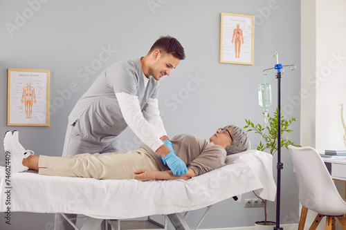 Sick senior patient lying on the couch in hospital receiving IV drip infusion. Young male nurse or doctor help elderly woman with vitamin injection therapy in medical clinic. Health care concept.