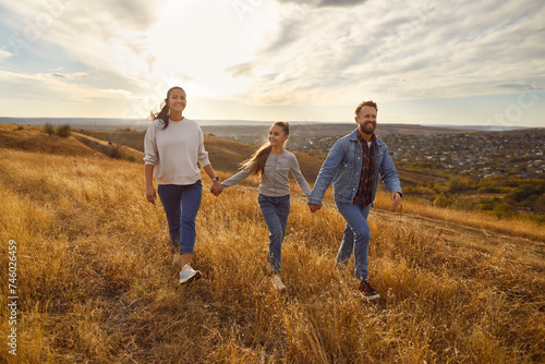 Happy smiling young family with child girl holding hands walking in the field enjoying nature at sunset. Mother, father and their daughter spending time outdoors. Family leisure concept.