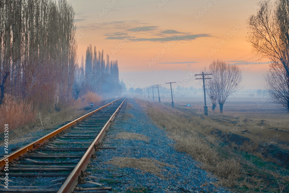 Fototapeta premium A railroad disappearing into the distance amidst fields in rural scenery, accompanied by old wooden electrical poles running alongside, against the backdrop of a sunset sky and distant light mist.