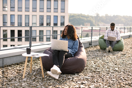 Successful business woman dressed in denim shirt and pants sitting at chair bag on roof top outdoors and typing on modern laptop, African American colleague works sitting in bag chair in background. © sofiko14