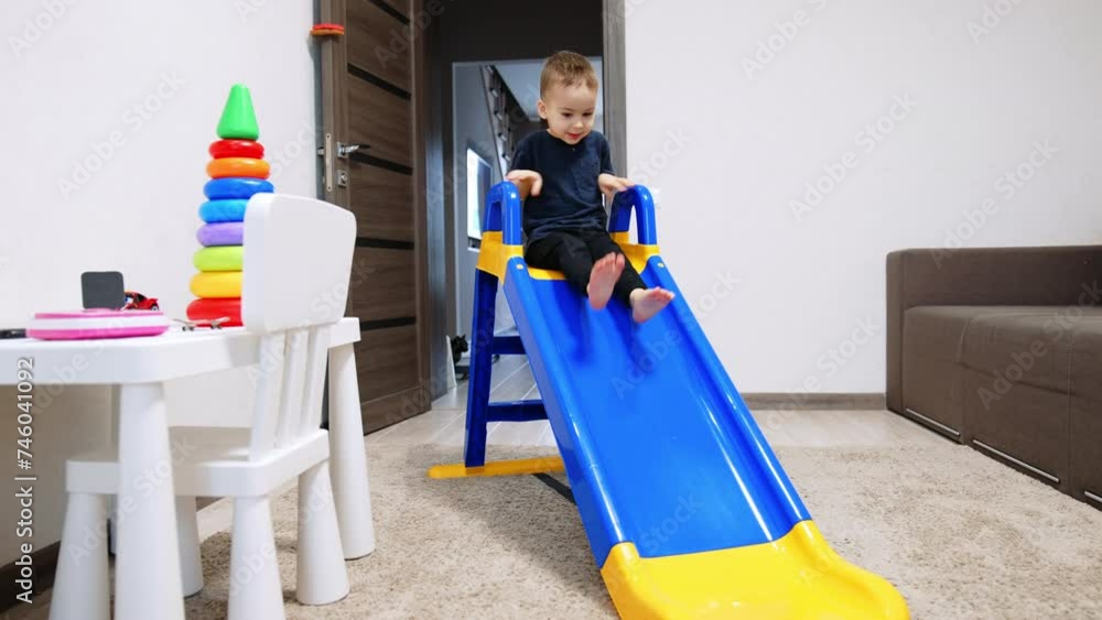 Smiling toddler in blue t-shirt and black pants barefoot sits on the slide. Baby boy slides down slowly. Kid playing at home. Smiling toddler in blue t-shirt and black pants barefoot sits on the slide. Baby boy slides down slowly. Kid playing at home.