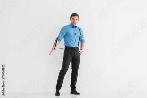 A man in a blue shirt, classic trousers, suspenders and a bow tie against a white wall in the studio. The man removes the suspender with one hand