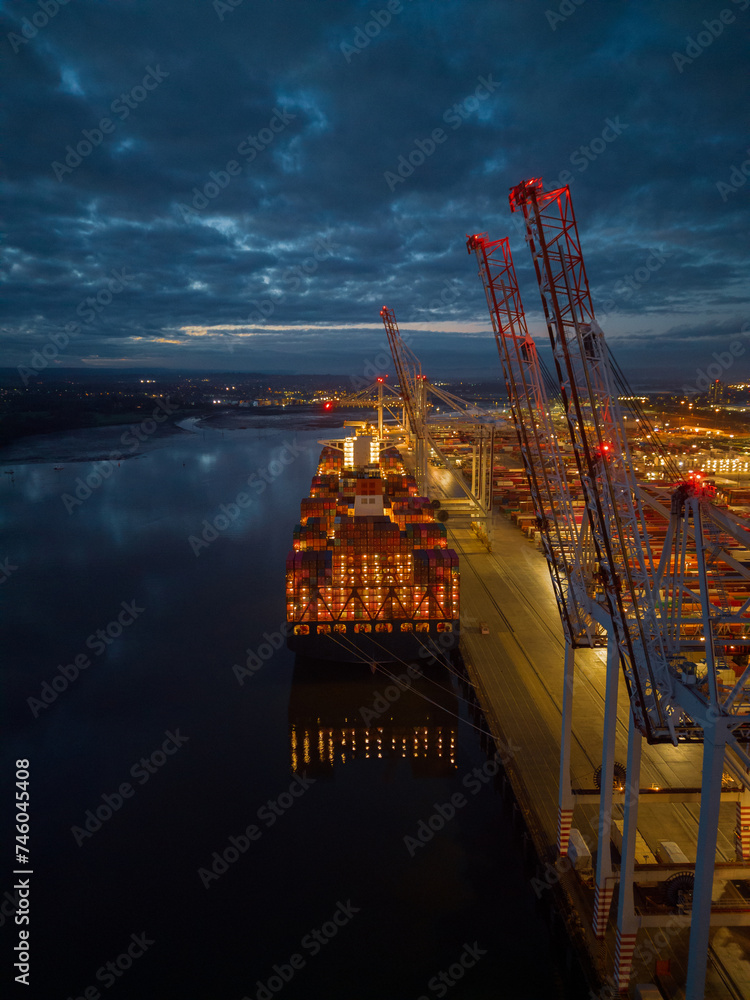 Beautiful container ship stern lights with reflection at dusk. Tall port cranes in Southampton Docks UK. Aerial vertical view. Calm cloudy night.