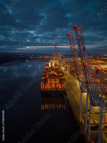 Obraz na płótnie Beautiful container ship stern lights with reflection at dusk. Tall port cranes in Southampton Docks UK. Aerial vertical view. Calm cloudy night. w salonie