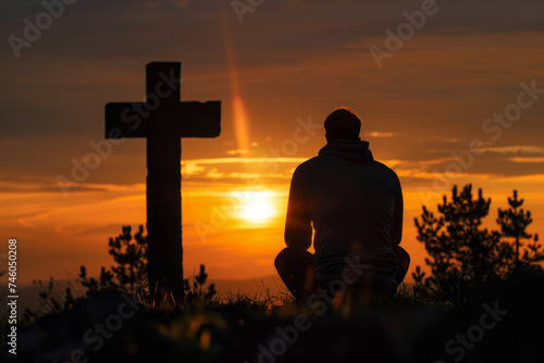 Silhouette of praying man at hilltop cross with warm glow of a sunset. Hope for salvation, request for help to heaven. Concept of faith in God
