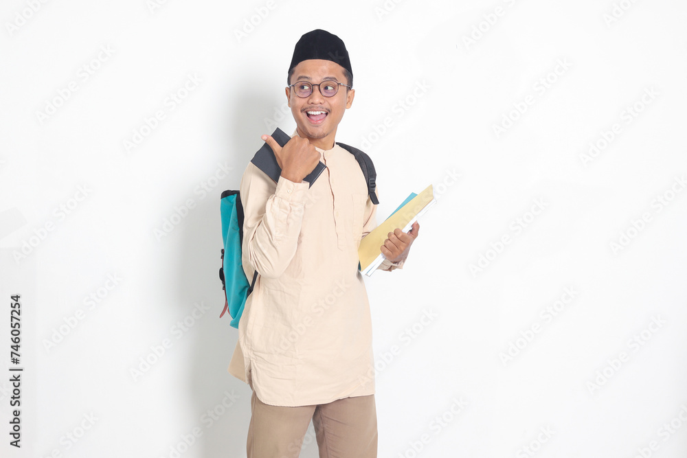 Portrait of excited student Asian muslim man in koko shirt with skullcap carrying backpack, holding school books, pointing to the side. Islamic education concept. Isolated image on white background Portrait of excited student Asian muslim man in koko shirt with skullcap carrying backpack, holding school books, pointing to the side. Islamic education concept. Isolated image on white background