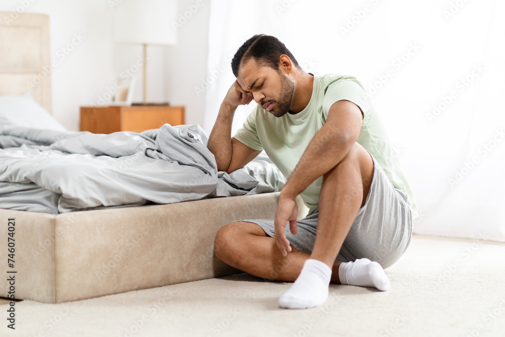 Unhappy millennial african american man sitting by bed at home