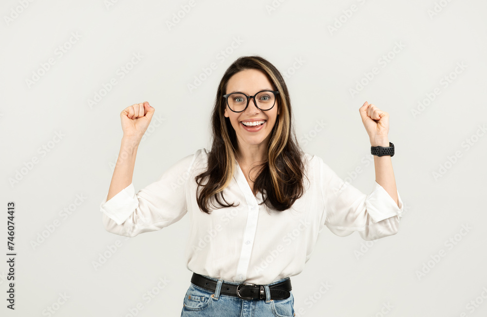 Joyful woman with glasses raising fists in excitement, wearing a white shirt and blue jeans Joyful woman with glasses raising fists in excitement, wearing a white shirt and blue jeans