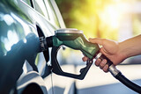 Close-up of a hand holding a green pump, petrol nozzle, refueling a car at a fuel station, with blurred natural light in the background