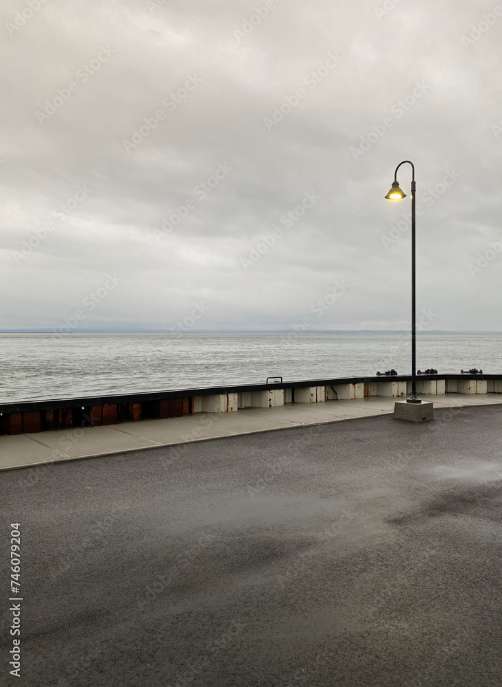 Lone lamppost at the end of a quay on a cloudy, gray day. Pier in the sea. A harbor bank and river waves in the distance on the horizon.