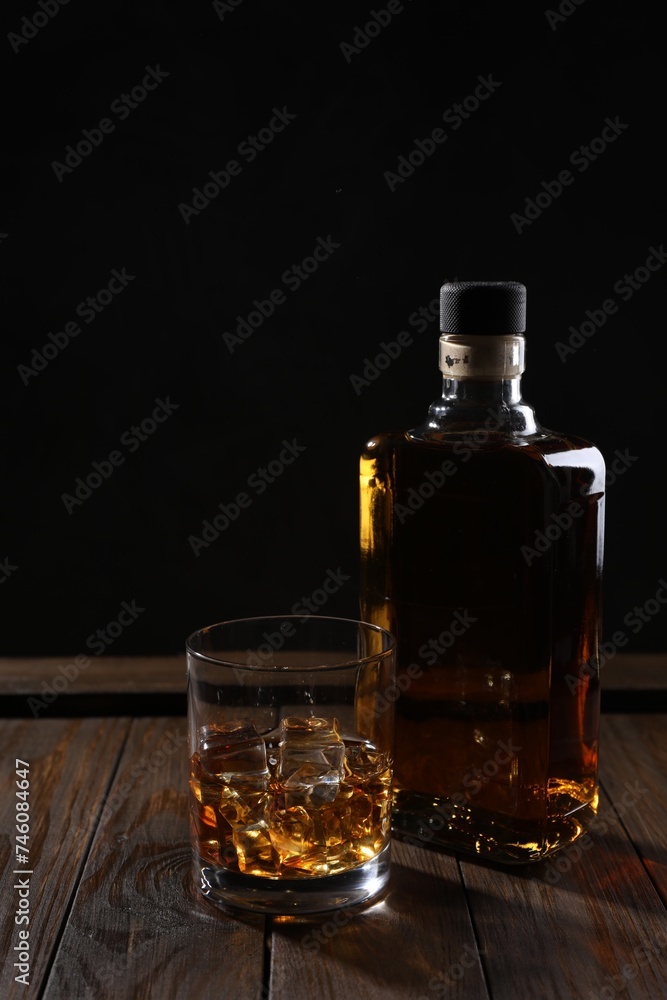 Whiskey with ice cubes in glass and bottle on wooden crate against black background Whiskey with ice cubes in glass and bottle on wooden crate against black background