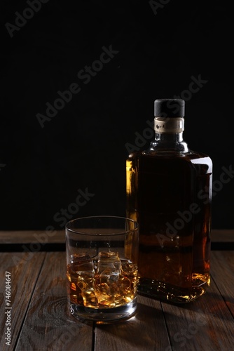 Whiskey with ice cubes in glass and bottle on wooden crate against black background
