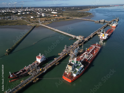 Multiple oil tankers moored at Fawley oil terminal. Aerial view of pipelines connected to biggest oil refinery in the UK with white fuel storage tanks. Southampton Docks at the distance.