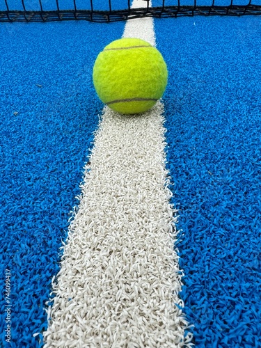 Close-up view of a line and texture of blue paddle tennis court and lonely yellow tennis ball. Artificial grass paddle court surface.