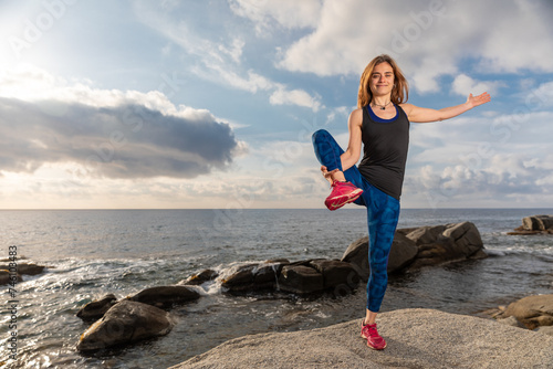 Young woman dressed in sports clothing, doing exercices with extended hand to right foot, on some rocks with the sea in the background photo
